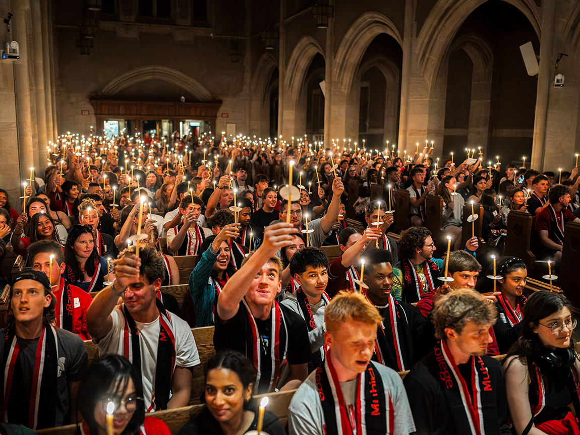 A large group of college students in a chapel raises candles high in the air